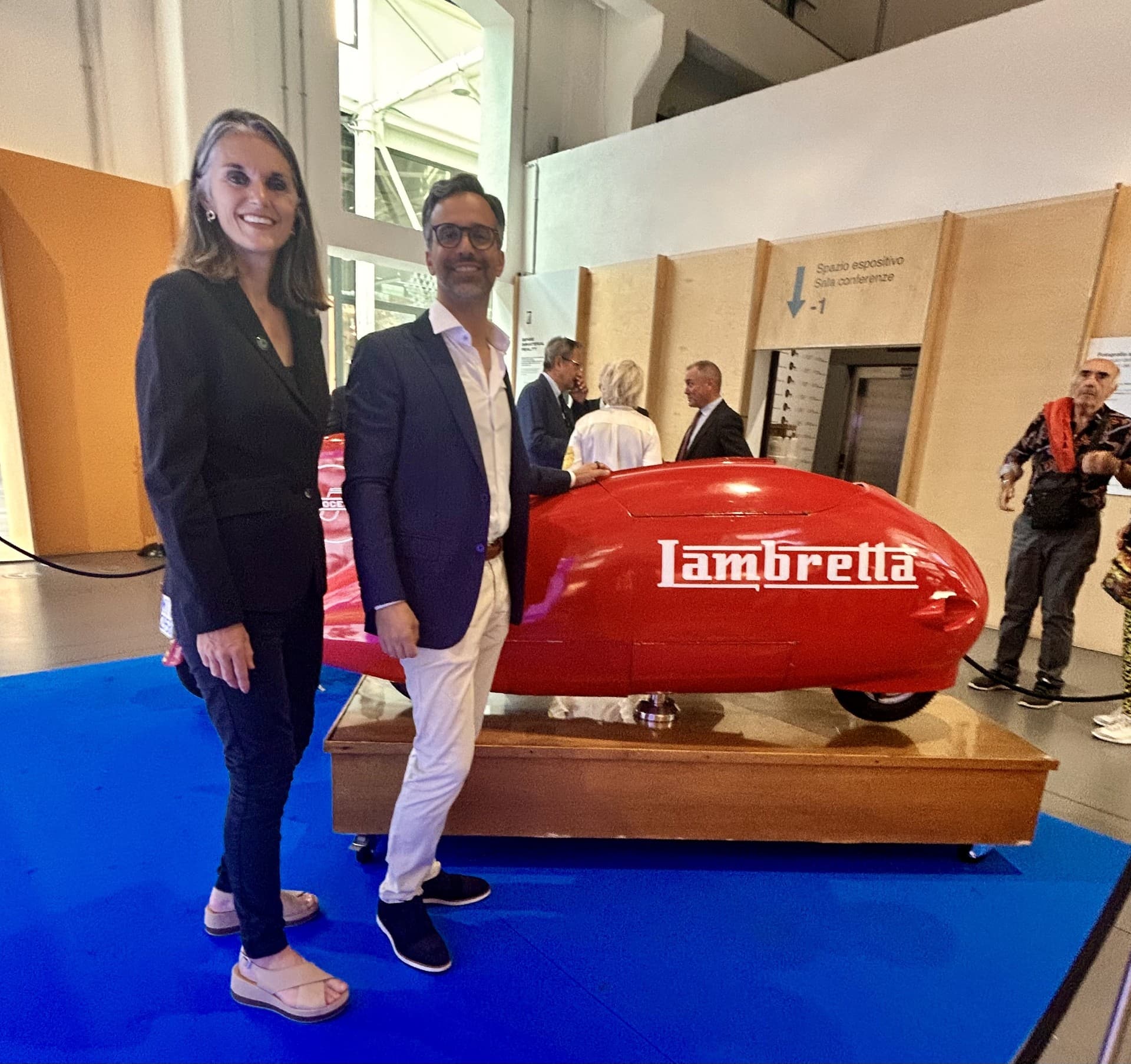 A woman wearing a black suit next to a man in navy blue blazer and chinos smiling proudly next to a small red vehicle